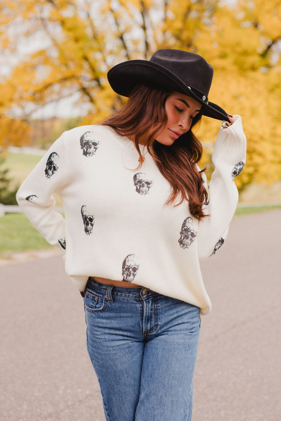 A person wearing a black cowboy hat and an Ivory Skull Sweater from Panache Apparel Collection featuring a black skull pattern.