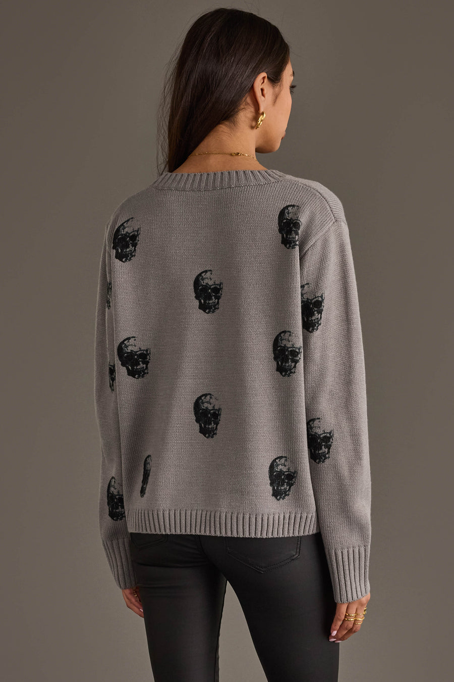 A person with long dark hair is seen from the back against a plain background, wearing the panacheaccessories Gray Skull Sweater, a relaxed fit gray knit sweater featuring black skull prints, paired with black pants.