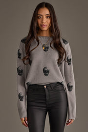 A woman with long brown hair models the panacheaccessories Gray Skull Sweater, a relaxed fit gray knit featuring bold black skull patterns, paired with black pants and gold jewelry, set against a plain, neutral background.