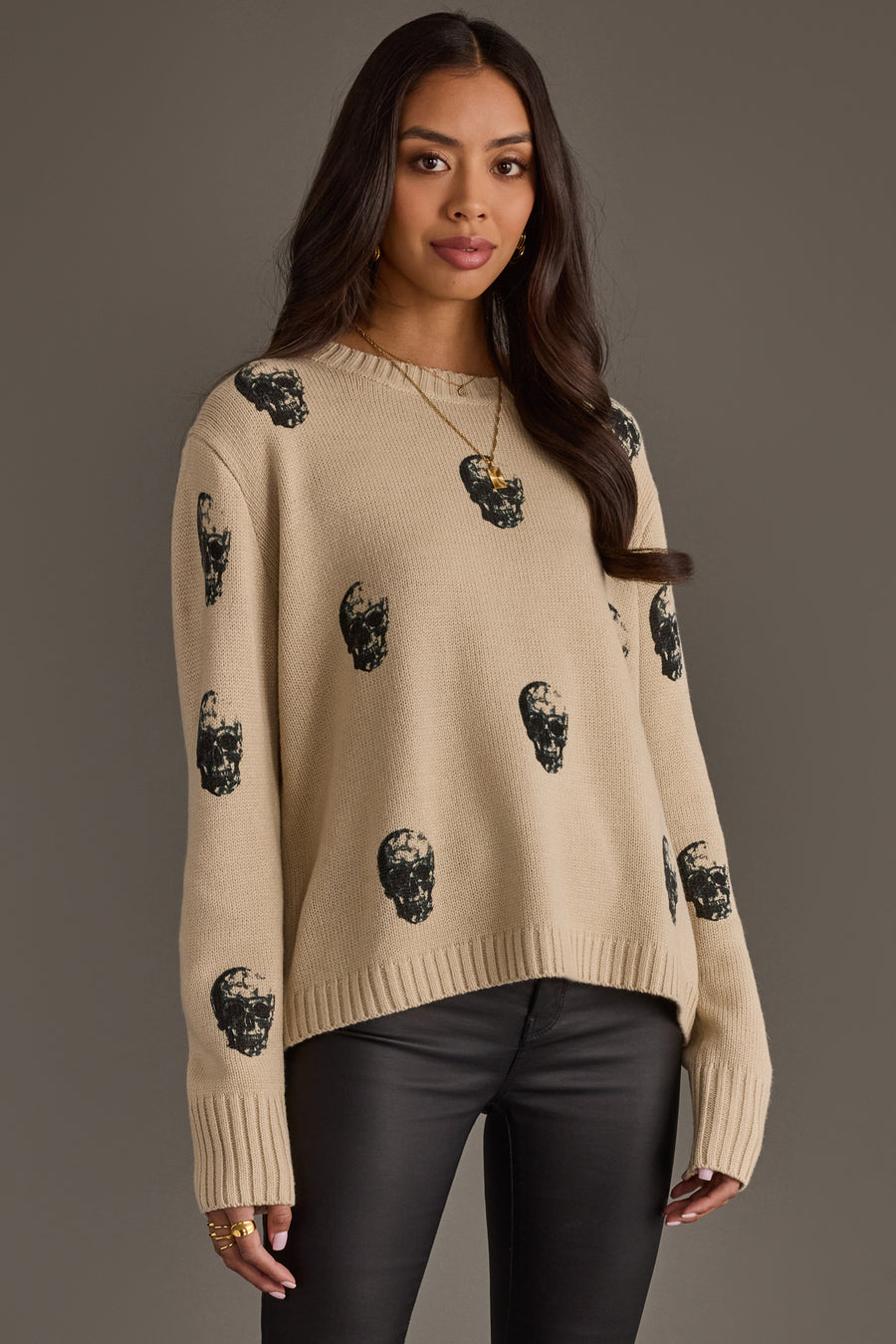 A woman with long brown hair wears the panacheaccessories Tan Skull Sweater, featuring a relaxed fit, paired with black leather pants and standing against a plain gray background.