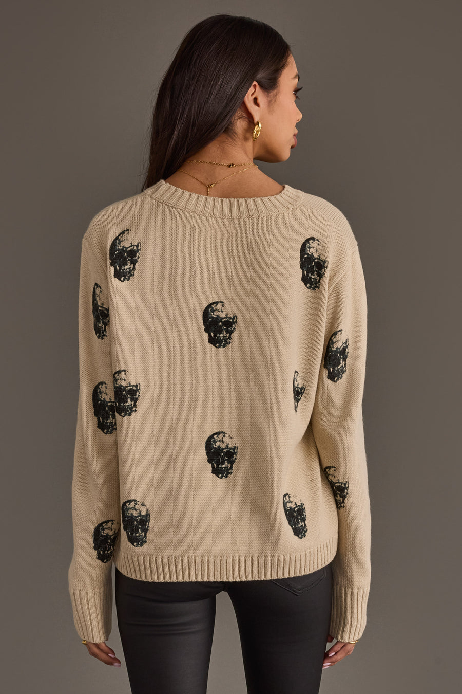 A woman with long dark hair, wearing the panacheaccessories Tan Skull Sweater in a relaxed fit paired with black pants, is standing with her back to the camera against a plain gray background.