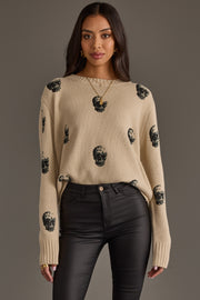 A woman wearing the panacheaccessories Tan Skull Sweater, featuring a relaxed fit and long sleeves, paired with black high-waisted pants. She stands against a plain background with loose hair and gold jewelry.