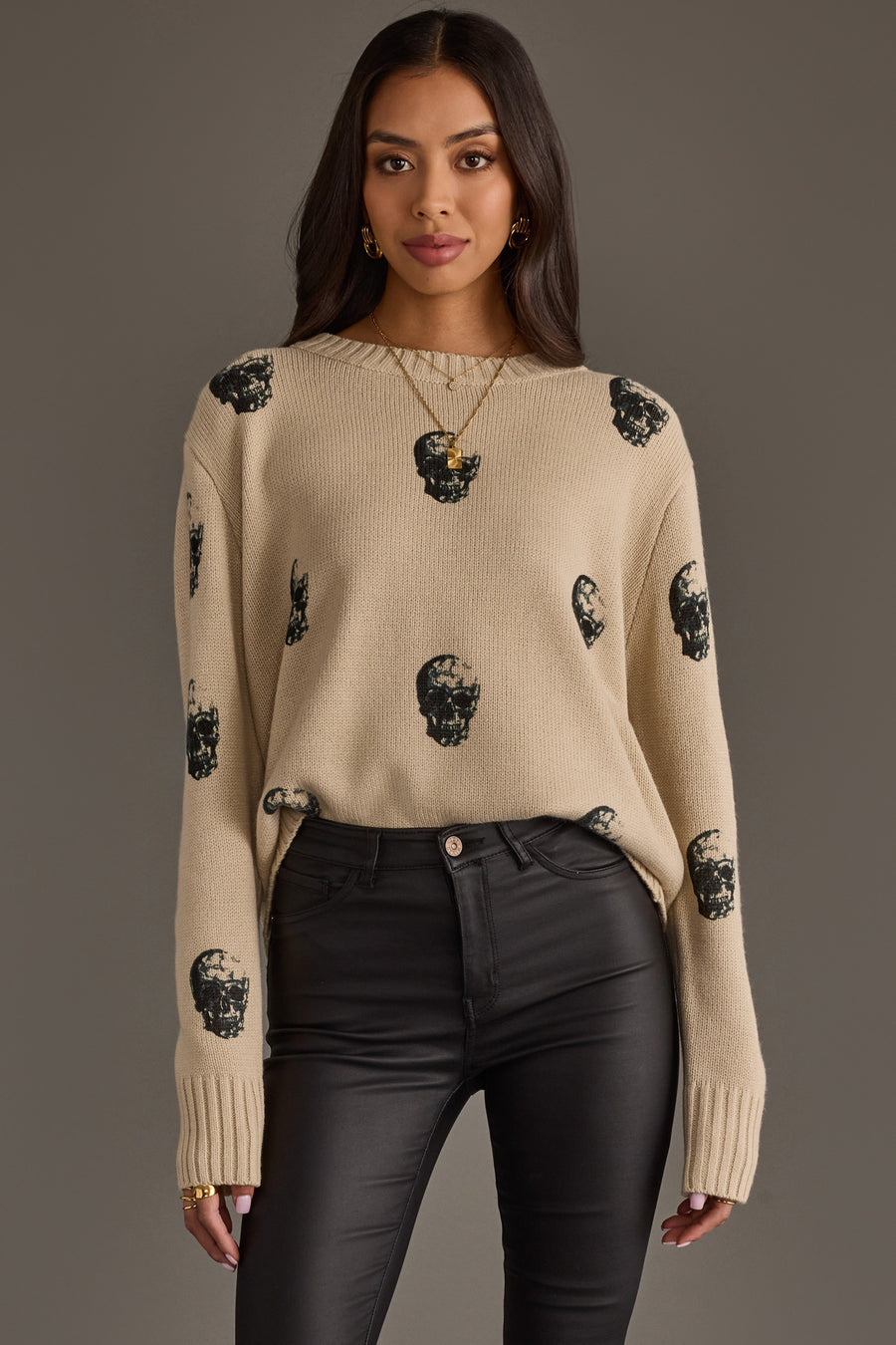 A woman wearing the panacheaccessories Tan Skull Sweater, featuring a relaxed fit and long sleeves, paired with black high-waisted pants. She stands against a plain background with loose hair and gold jewelry.