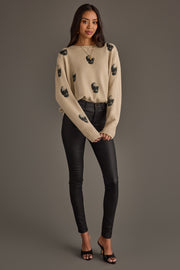 A woman stands against a gray background, wearing the panacheaccessories Tan Skull Sweater, a tan knit top adorned with black beaded embellishments, paired with black fitted pants and black open-toe heels. Her long, dark hair cascades over her shoulders, and her hands are gently clasped in front of her.