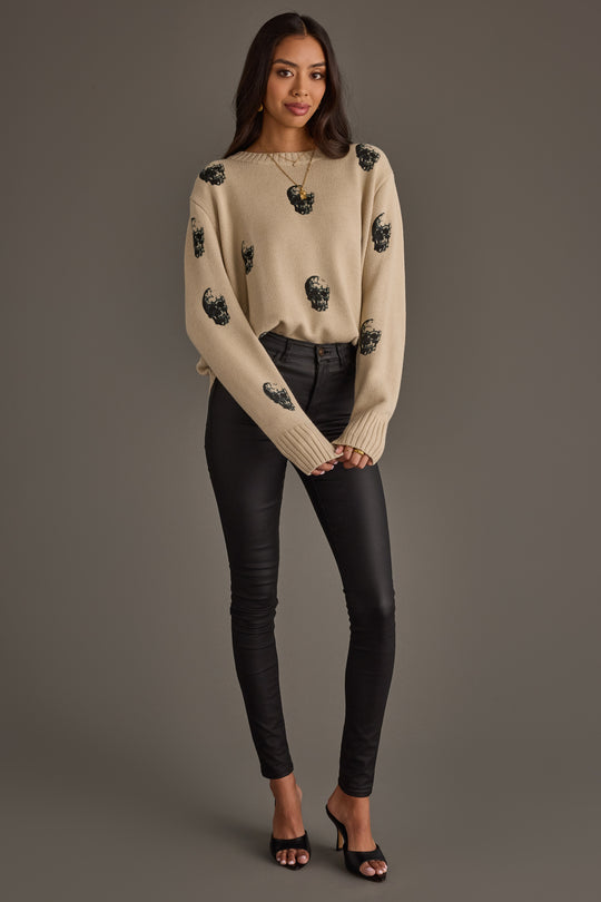 A woman stands against a gray background, wearing the panacheaccessories Tan Skull Sweater, a tan knit top adorned with black beaded embellishments, paired with black fitted pants and black open-toe heels. Her long, dark hair cascades over her shoulders, and her hands are gently clasped in front of her.