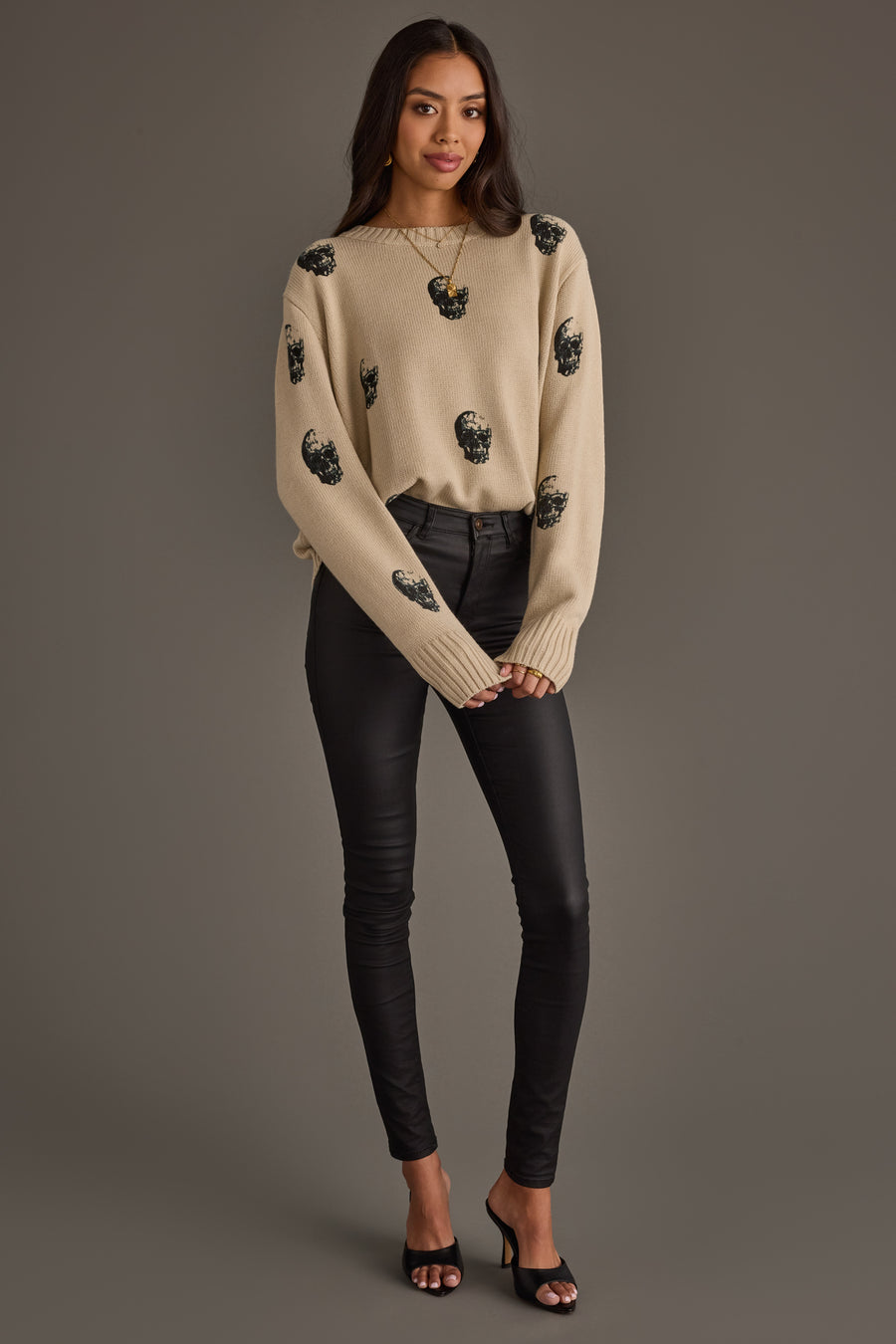 A woman stands against a gray background, wearing the panacheaccessories Tan Skull Sweater, a tan knit top adorned with black beaded embellishments, paired with black fitted pants and black open-toe heels. Her long, dark hair cascades over her shoulders, and her hands are gently clasped in front of her.
