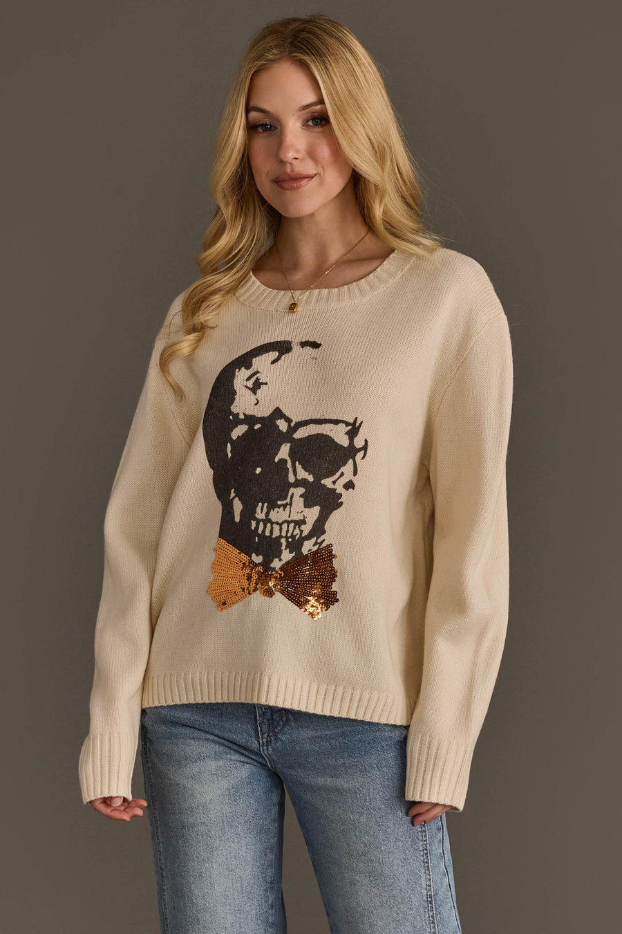 A woman models the Panache Apparel Collection’s Large Skull Sweater with Bow Tie, a cream sweater featuring a bold black skull graphic and shimmering gold sequin bow tie, paired with blue jeans. She has long blonde hair and poses against a plain gray background.