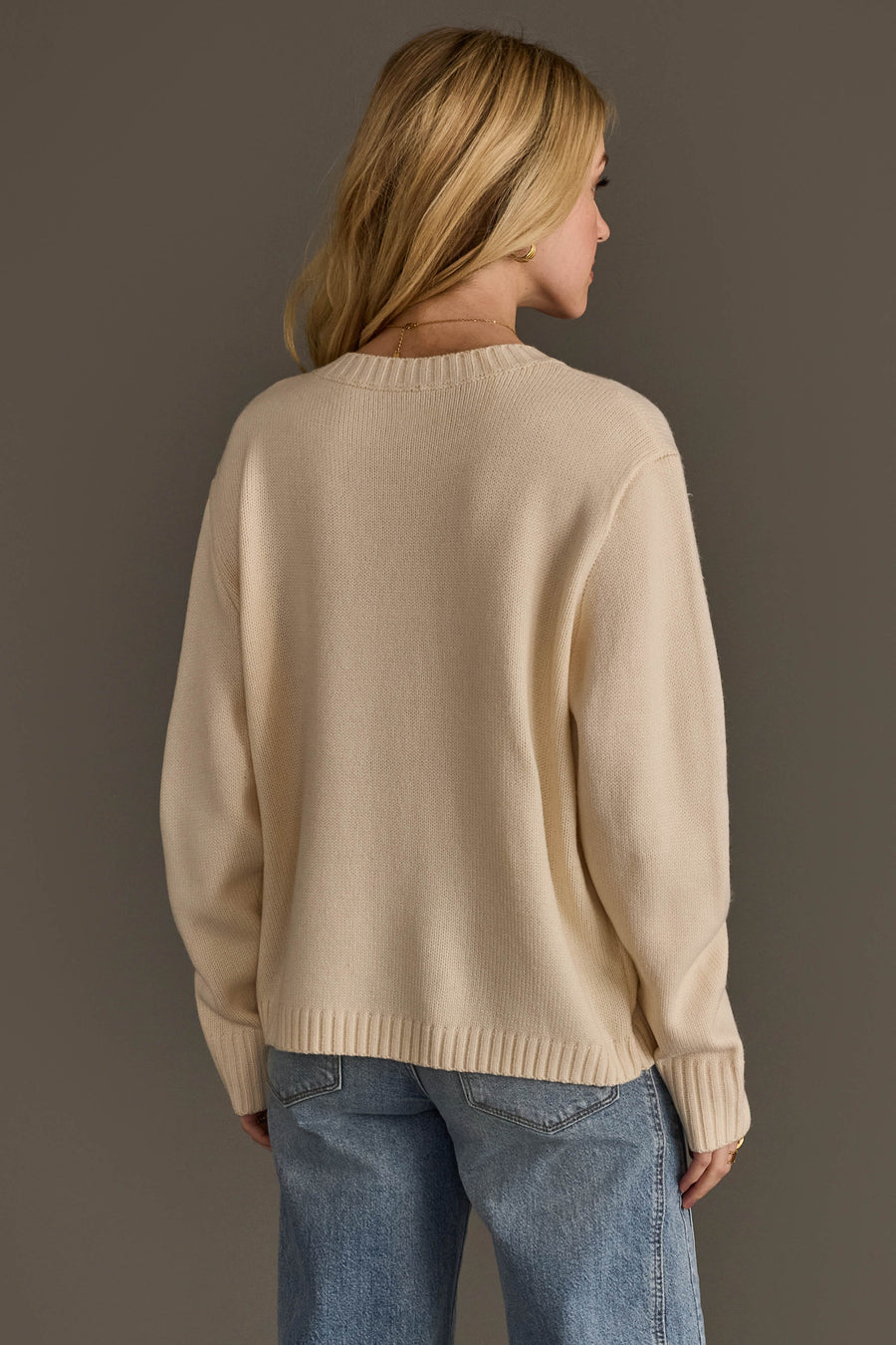 A person with long blonde hair wears the Panache Apparel Collection’s Large Skull Sweater with Bow Tie in a cream color, paired with light blue jeans, facing away from the camera against a plain gray background.