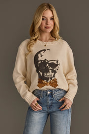 A woman with long blonde hair is wearing the Panache Apparel Collection Large Skull Sweater with Bow Tie, a cream sweater featuring a sequined skull and bowtie design, paired with blue jeans and standing against a plain gray background.