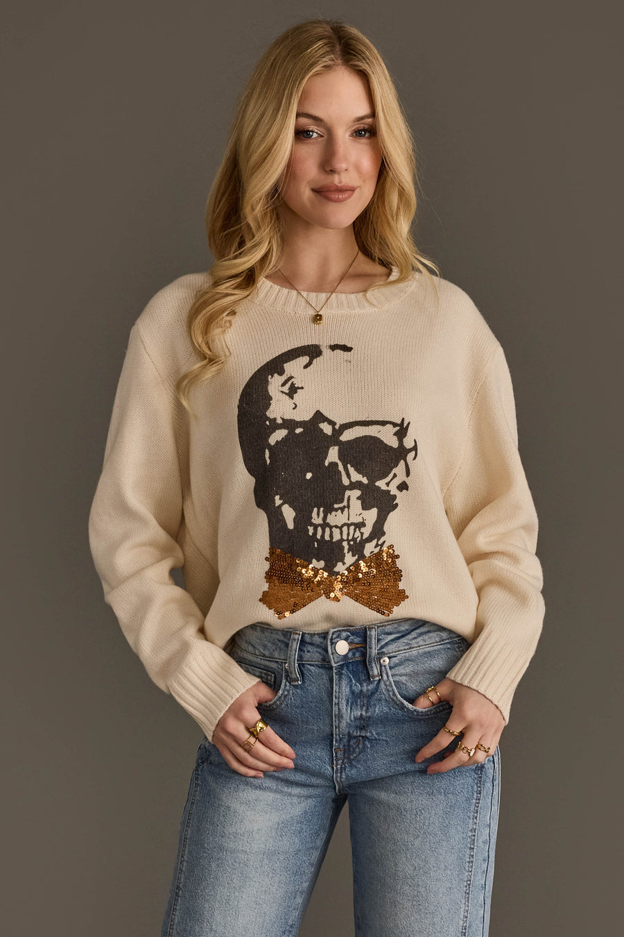 A woman with long blonde hair is wearing the Panache Apparel Collection Large Skull Sweater with Bow Tie, a cream sweater featuring a sequined skull and bowtie design, paired with blue jeans and standing against a plain gray background.