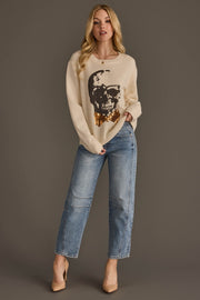 A woman stands against a gray background wearing the Panache Apparel Collection’s Large Skull Sweater with Bow Tie, a beige sweater featuring a sequin skull graphic, paired with light blue jeans and nude heels. Her long blonde hair frames her face as she strikes a relaxed, confident pose with one hand in her pocket.