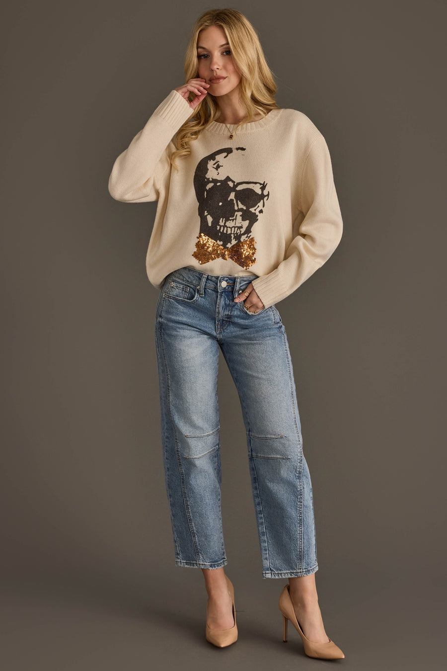 A woman stands against a gray background wearing the Panache Apparel Collection’s Large Skull Sweater with Bow Tie, paired with light blue jeans and beige heels.