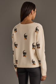 A woman with long dark hair, wearing large hoop earrings, a cream Skull Sweater with Bow Ties by Panache Apparel Collection, and black pants stands facing away from the camera against a plain grey background.