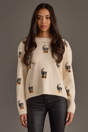 A woman with long dark hair wears the Skull Sweater with Bow Ties from Panache Apparel Collection and black leather pants, standing against a plain gray background.