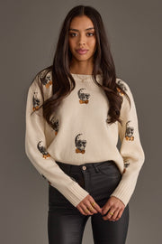 A woman with long dark hair wears the Panache Apparel Collection’s Skull Sweater with Bow Ties, featuring cream fabric adorned with black skull faces and orange bows, paired with black pants, standing against a plain gray background.
