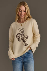 A smiling woman with long blonde hair models the Panache Apparel Collection’s Cowboy Sweater, featuring a lassoing cowboy on horseback design, paired with blue jeans and set against a plain gray background.