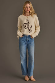 A person wearing the Cowboy Sweater by Panache Apparel Collection, paired with blue jeans and beige heels, stands against a plain brown background with hands in pockets and a slight smile.