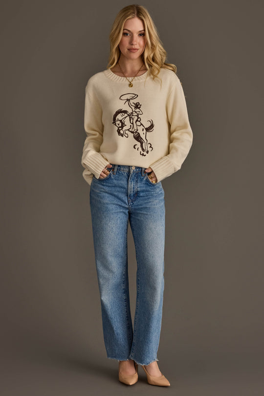 A person wearing the Cowboy Sweater by Panache Apparel Collection, paired with blue jeans and beige heels, stands against a plain brown background with hands in pockets and a slight smile.