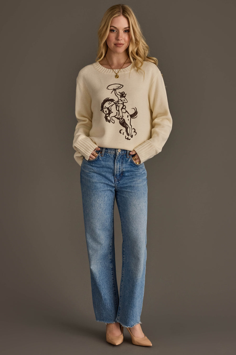 A person wearing the Cowboy Sweater by Panache Apparel Collection, paired with blue jeans and beige heels, stands against a plain brown background with hands in pockets and a slight smile.
