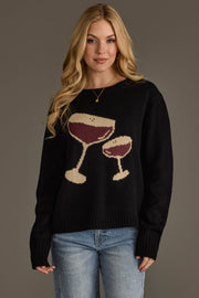 A woman with long blonde hair wears the Panache Apparel Collection’s Espresso Martini Sweater, a black knit top featuring two wine glasses, paired with light blue jeans as she stands against a plain gray background. The relaxed fit of the sweater adds effortless style to her look.