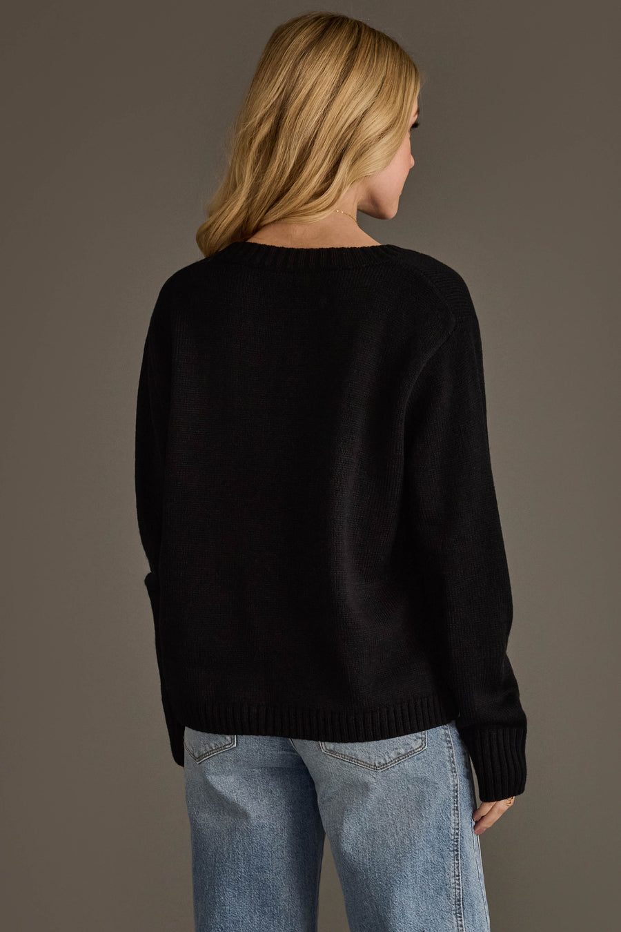 A person with long blonde hair is seen from behind, wearing the Panache Apparel Collection’s Espresso Martini Sweater—a relaxed fit black knit—paired with blue jeans, standing against a plain gray background.