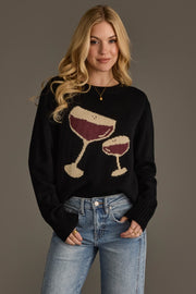A woman with long blonde hair wears the Panache Apparel Collection Espresso Martini Sweater, featuring two wine glass graphics, paired with light blue jeans and standing against a plain gray background.