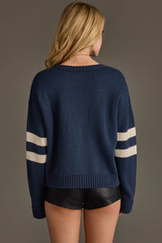 A woman with long blonde hair stands with her back to the camera against a plain background, wearing the Panache Apparel Collection Navy & White 89 Sweater paired with black leather shorts.