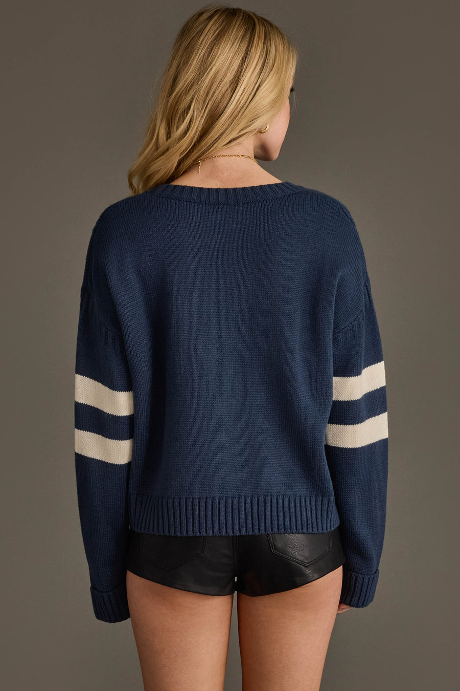 A woman with long blonde hair stands with her back to the camera against a plain background, wearing the Panache Apparel Collection Navy & White 89 Sweater paired with black leather shorts.