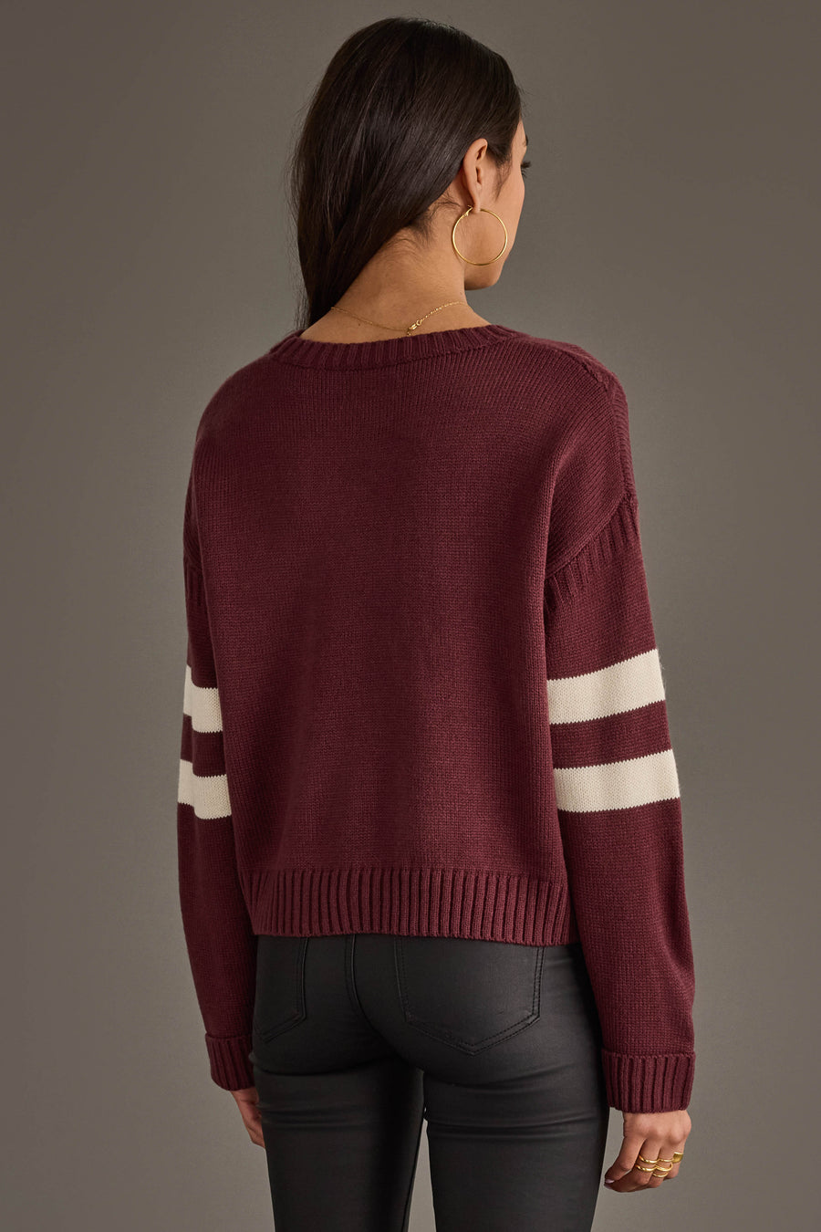 A woman with long dark hair, wearing gold hoop earrings and black pants, stands facing away from the camera against a plain background, showcasing the Panache Apparel Collection Burgundy & White 17 Sweater.