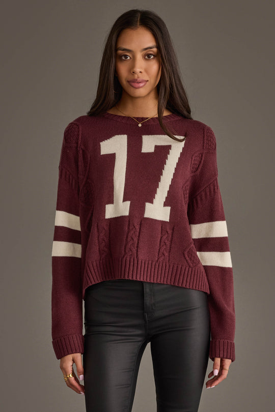 A woman models the Panache Apparel Collection's Burgundy & White 17 Sweater, featuring white sleeve stripes and a bold number 17 on the front, paired with black pants against a plain gray background.