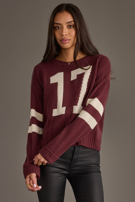 A woman with long brown hair models the Panache Apparel Collection's Burgundy & White 17 Sweater, featuring white sleeve stripes and the number 17, paired with black pants against a plain gray background.