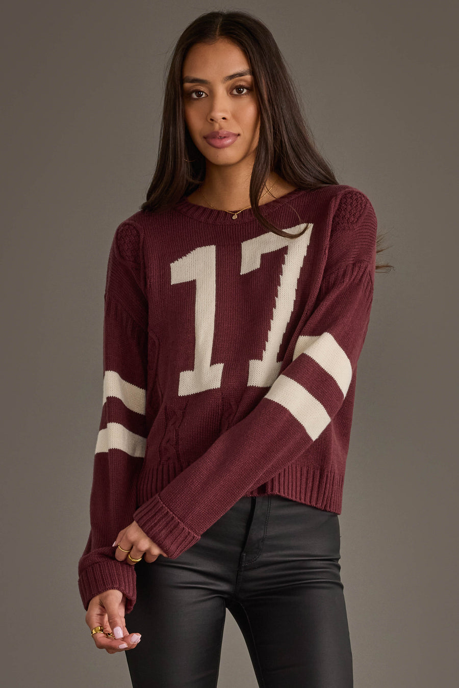 A woman with long brown hair models the Panache Apparel Collection's Burgundy & White 17 Sweater, featuring white sleeve stripes and the number 17, paired with black pants against a plain gray background.