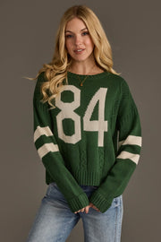 A woman with long blonde hair models the Panache Apparel Collection Dark Green & White 84 Sweater, featuring white stripes on the sleeves and the number 84 on the front, paired with blue jeans and set against a plain gray background.