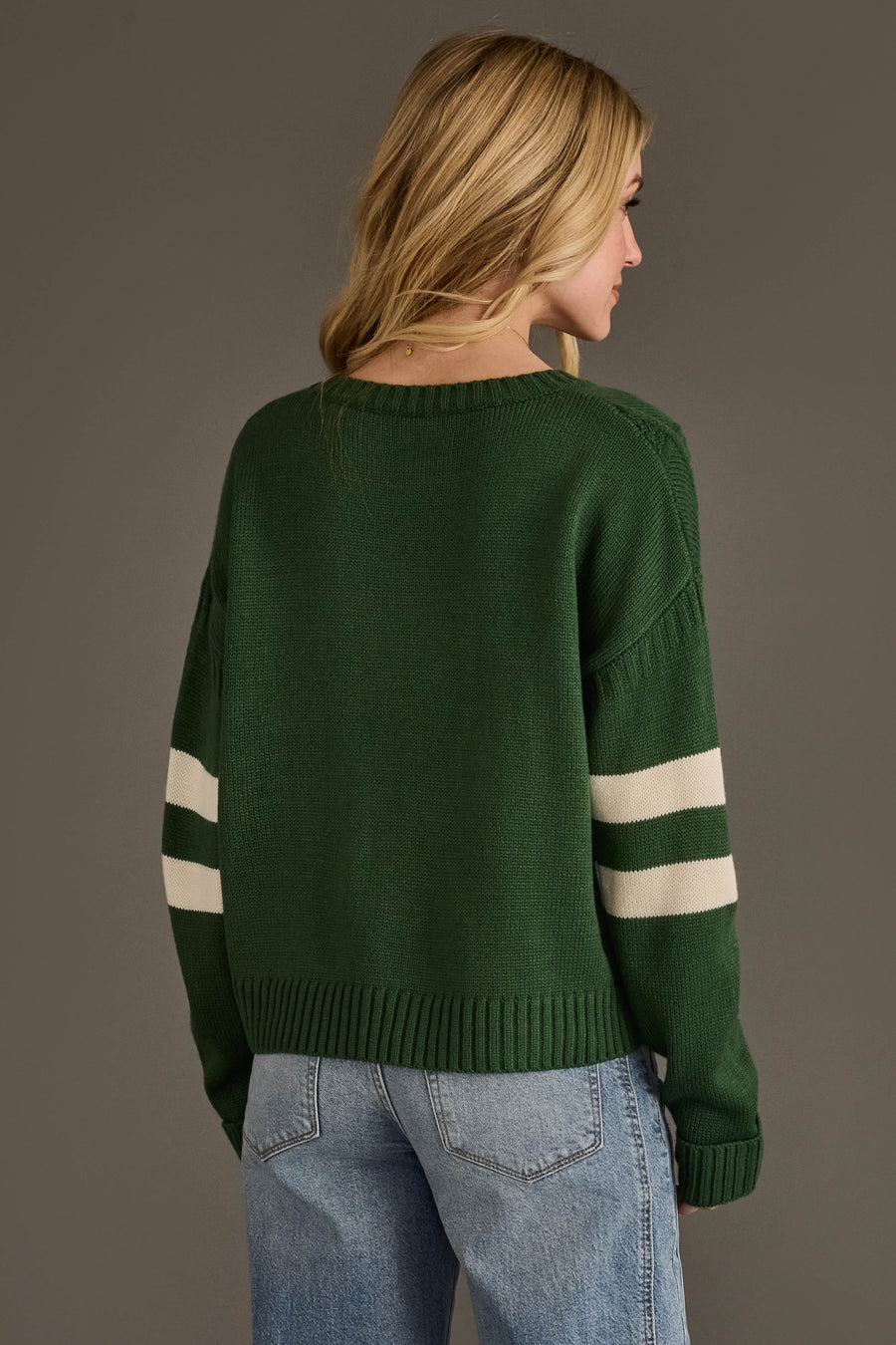 Wearing the Panache Apparel Collection’s Dark Green & White 84 Sweater featuring two white stripes on the sleeves, a person with long blonde hair and light blue jeans stands facing away from the camera against a plain background.