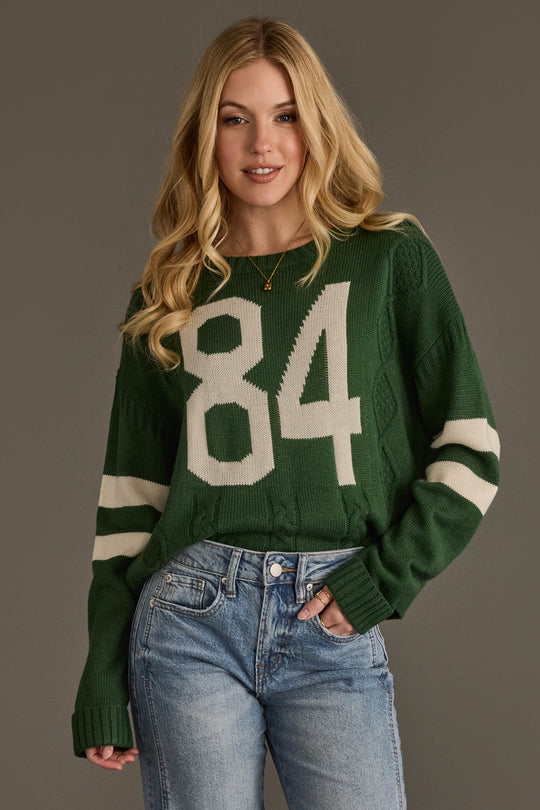 A person with long blonde hair wears the Panache Apparel Collection’s Dark Green & White 84 Sweater, featuring white stripes on the sleeves, paired with blue jeans. The background is plain gray.
