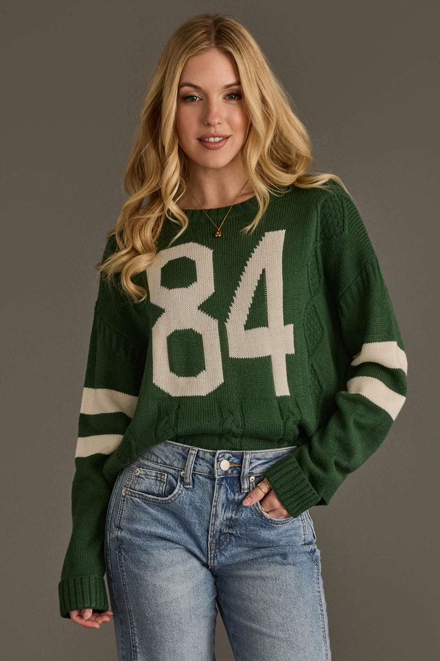A person with long blonde hair wears the Panache Apparel Collection’s Dark Green & White 84 Sweater, featuring white stripes on the sleeves, paired with blue jeans. The background is plain gray.