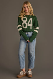 A woman with long blonde hair wears the Panache Apparel Collection Dark Green & White 84 Sweater, paired with blue cuffed jeans and brown sneakers, standing against a plain gray background.