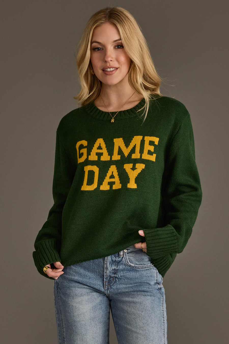 A woman stands against a plain gray background, smiling with one hand in her pocket while wearing the Green & Gold Game Day Sweater from Panache Apparel Collection, paired with blue jeans and a necklace.