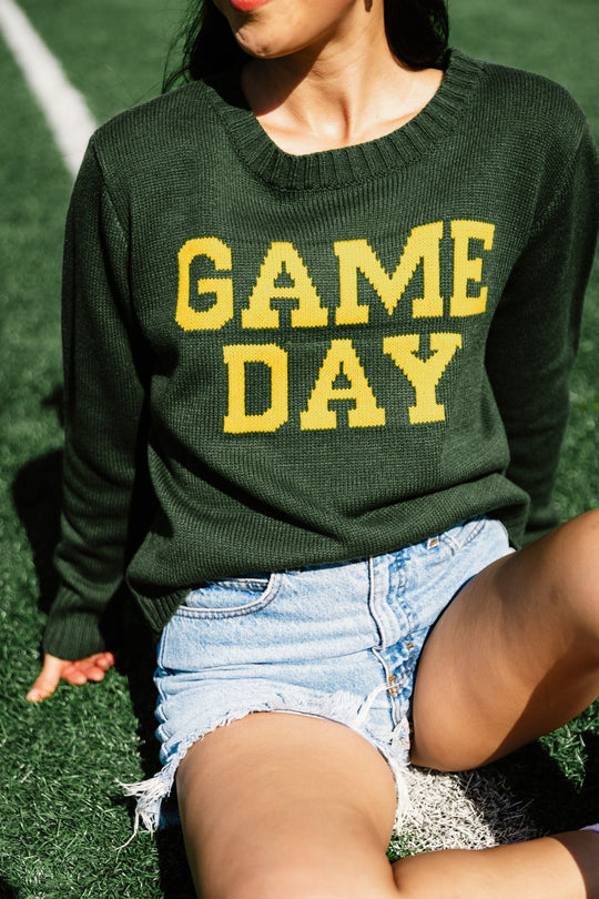 A person wearing the Panache Apparel Collection’s Green & Gold Game Day Sweater—a relaxed fit green knit sweater with “GAME DAY” in bold yellow letters—paired with denim shorts, sits on the grass, likely on a sports field.
