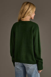 A person with long blonde hair, seen from behind, is wearing the Panache Apparel Collection’s Green & Gold Game Day Sweater paired with light blue jeans, standing against a plain brown background.