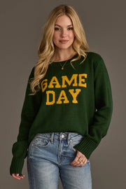 A woman with long blonde hair wears the Green & Gold Game Day Sweater by Panache Apparel Collection, featuring yellow "GAME DAY" lettering, paired with blue jeans as she stands against a neutral gray background.