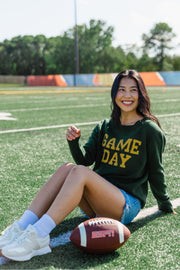 A woman in a relaxed fit Green & Gold Game Day Sweater from Panache Apparel Collection sits smiling on a football field next to an ESPN football, with trees and a colorful fence in the background.