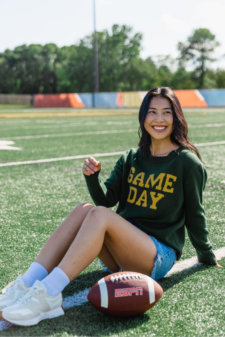 A woman in a relaxed fit Green & Gold Game Day Sweater from Panache Apparel Collection sits smiling on a football field next to an ESPN football, with trees and a colorful fence in the background.