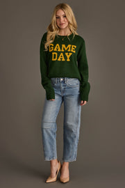 A woman with long blonde hair is standing against a gray background, wearing the Panache Apparel Collection Green & Gold Game Day Sweater featuring yellow "GAME DAY" lettering. She pairs it with light blue jeans and nude heels, and has a relaxed, smiling expression.