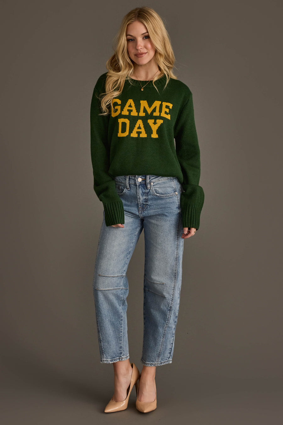 A woman with long blonde hair is standing against a gray background, wearing the Panache Apparel Collection Green & Gold Game Day Sweater featuring yellow "GAME DAY" lettering. She pairs it with light blue jeans and nude heels, and has a relaxed, smiling expression.