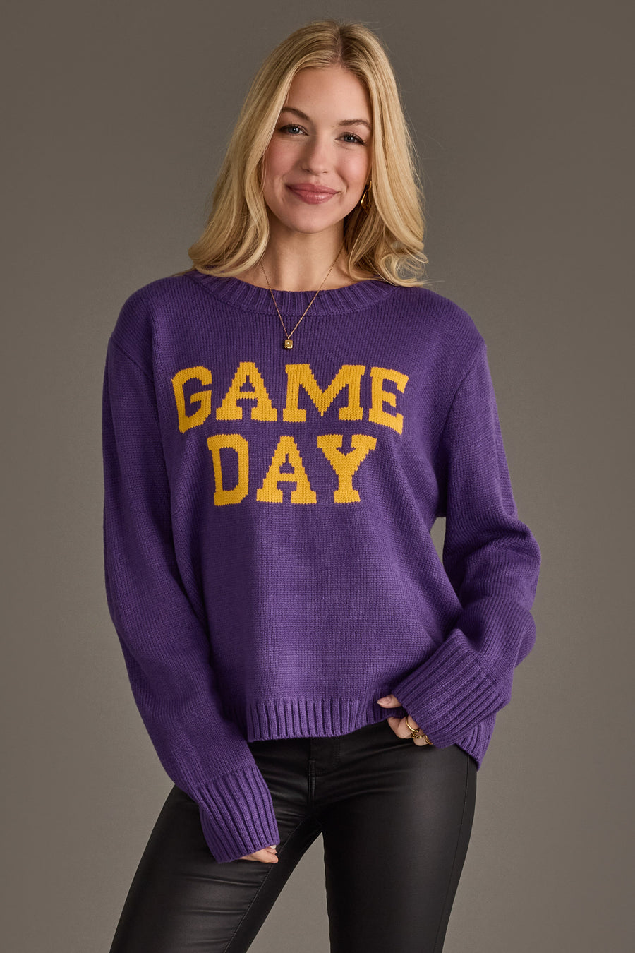 A woman models the Panache Apparel Collection's Purple & Gold Game Day Sweater, featuring bold yellow "GAME DAY" text, paired with black pants and a necklace while smiling at the camera against a plain gray background.