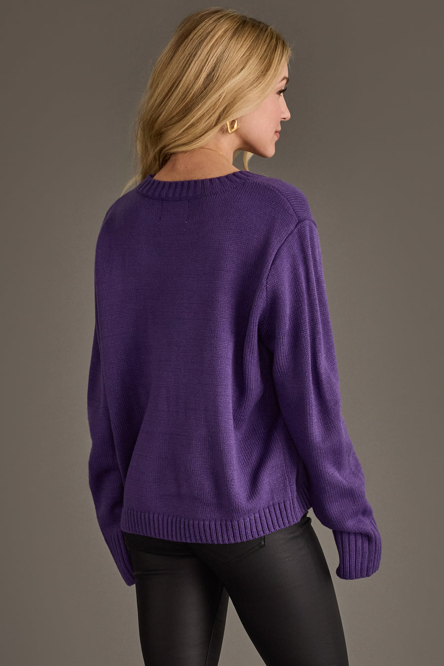 A woman with blonde hair, wearing gold hoop earrings and the Panache Apparel Collection’s Purple & Gold Game Day Sweater paired with black pants, stands facing away from the camera against a plain gray background.