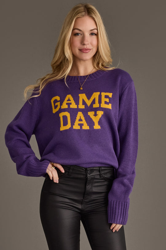A woman with long blonde hair stands and smiles against a plain background, wearing the Panache Apparel Collection’s Purple & Gold Game Day Sweater paired with black leather pants.