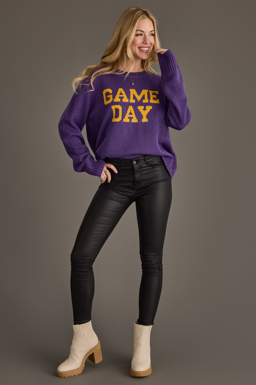 A woman smiles while standing with one hand on her hip, wearing the Panache Apparel Collection Purple & Gold Game Day Sweater, paired with black pants and beige boots against a plain gray background.