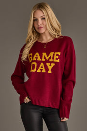 A woman with long blonde hair is wearing the Panache Apparel Collection’s Maroon & Gold Game Day Sweater, styled with black leather pants, and standing against a plain gray background.